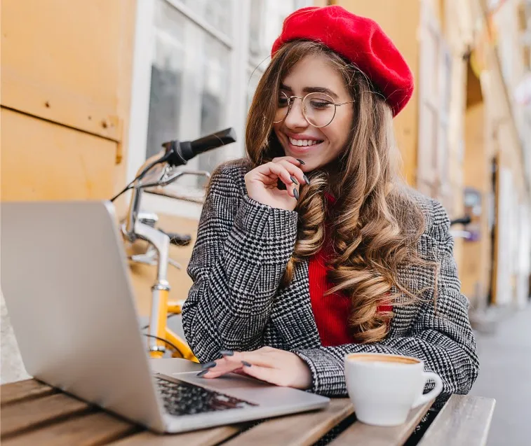 Moça de boina vermelha sorrindo diante do notebook, com uma xícara de café